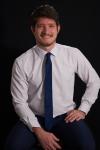 Jacob sits in a white shirt and blue tie in front of a black background. 