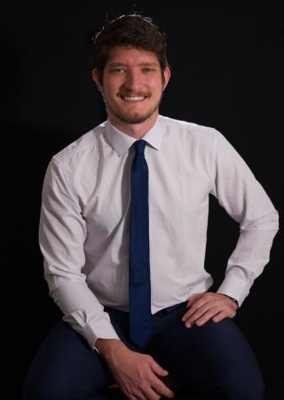 Jacob sits in a white shirt and blue tie in front of a black background.