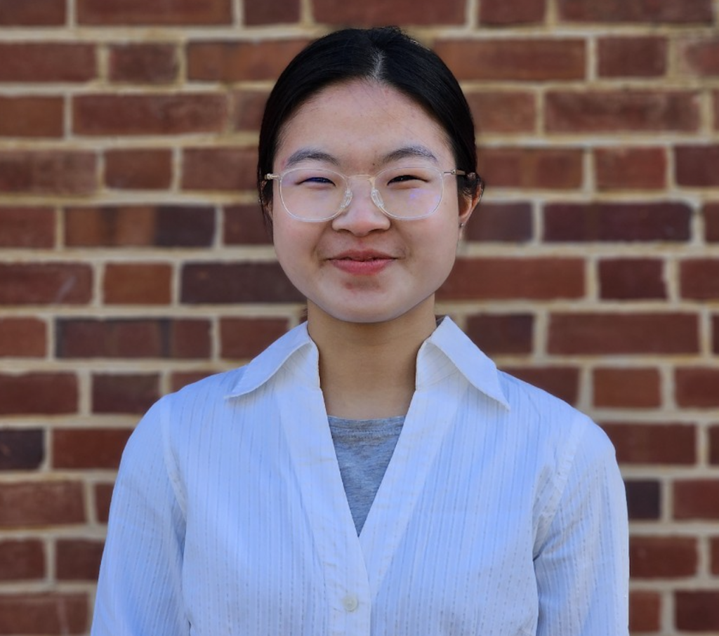 Hana, wearing glasses, stares into the camera with a brick background. 