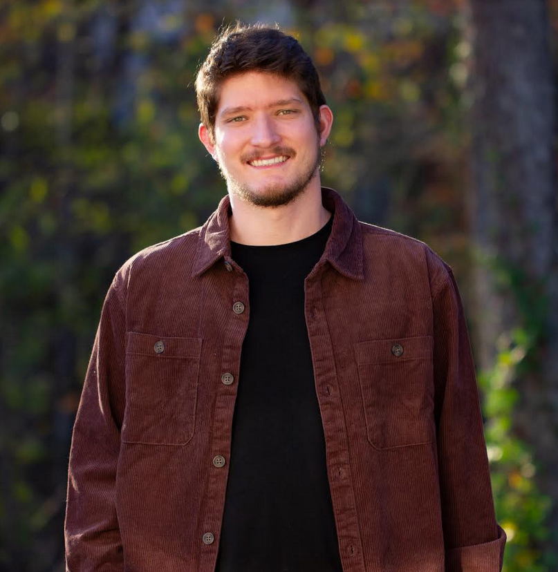 Jacob stands in a brown jacket against a forest backdrop.