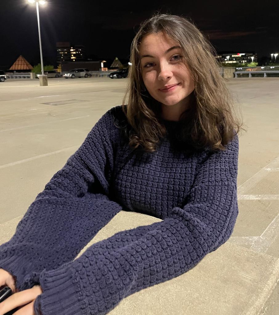 Katy sits in at a concrete table in a parking lot wearing a sweater