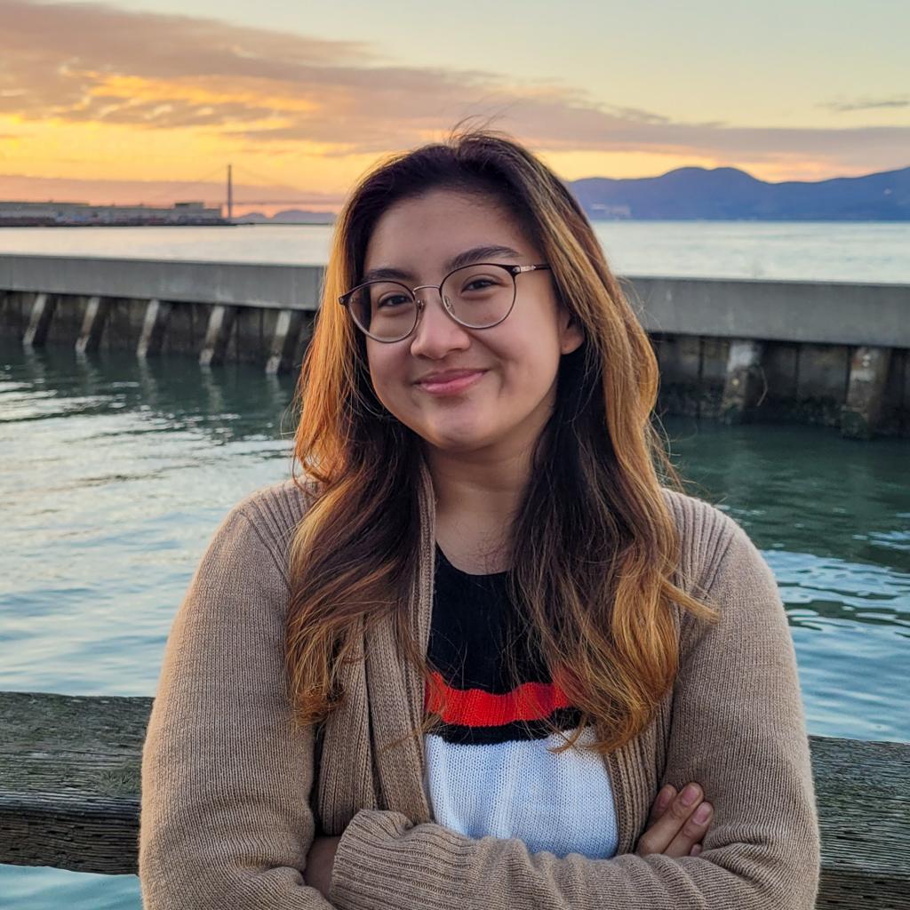 Irene,  wearing glasses, stands with her arms crossed in front of a pier. 